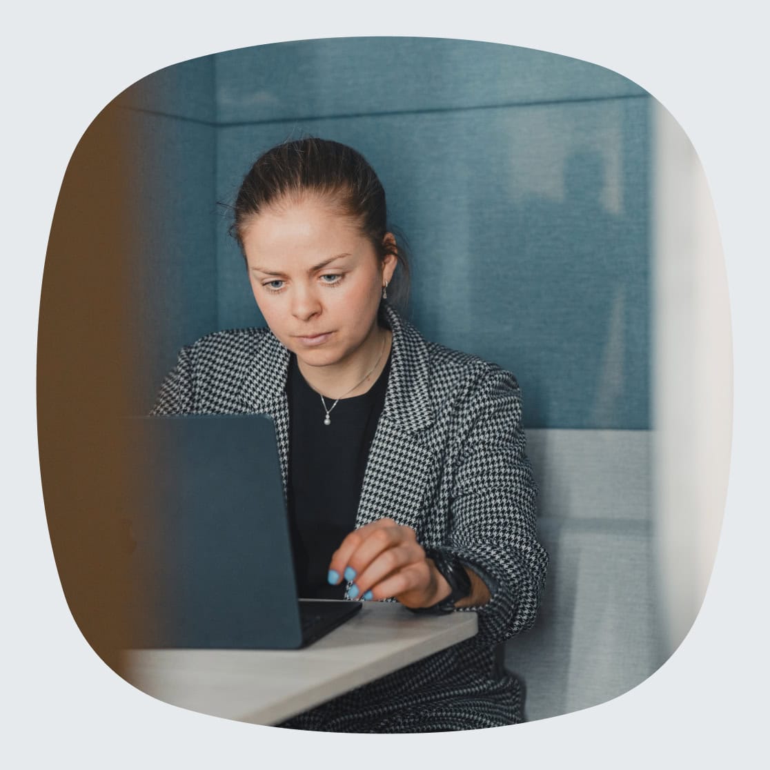 En kvinne jobber fokusert på en bærbar datamaskin i en arbeidskrok med blå polstrede vegger. Hun har på seg en rutete blazer og ser konsentrert på skjermen.