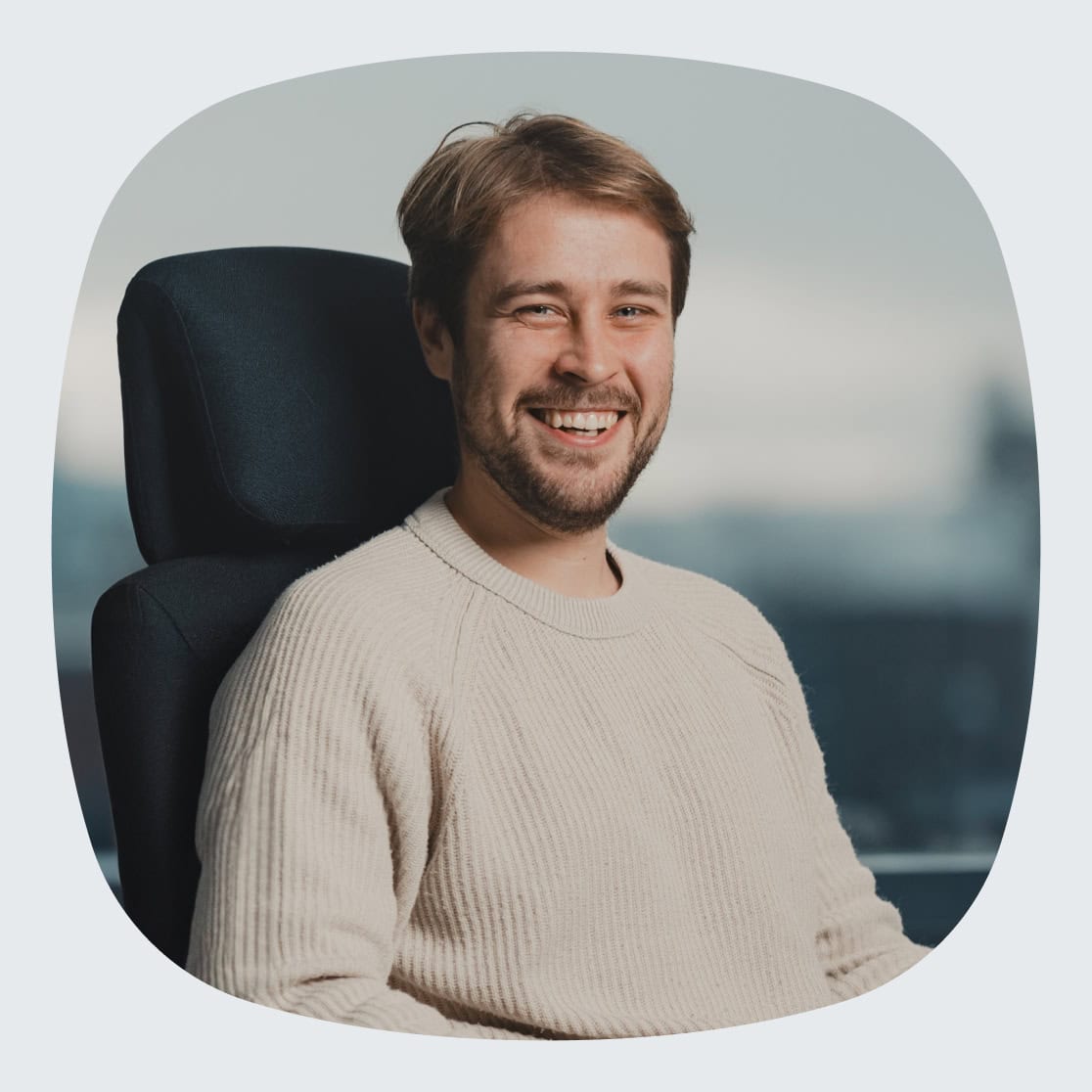 En ung mann med kort, lyst hår og skjegg, iført en beige genser, smiler mens han sitter i en kontorstol foran et stort vindu med utsikt over Oslo.