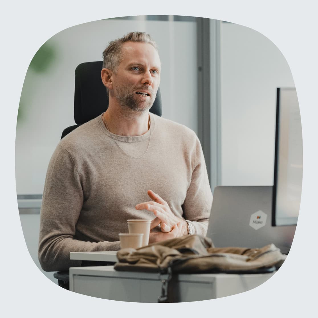 En mann med kort hår, skjegg og beige genser sitter ved et skrivebord. På pulten står en kaffekopp, en veske og en bærbar datamaskin med en «Make»-logo. Bakgrunnen viser store vinduer som slipper inn naturlig lys.