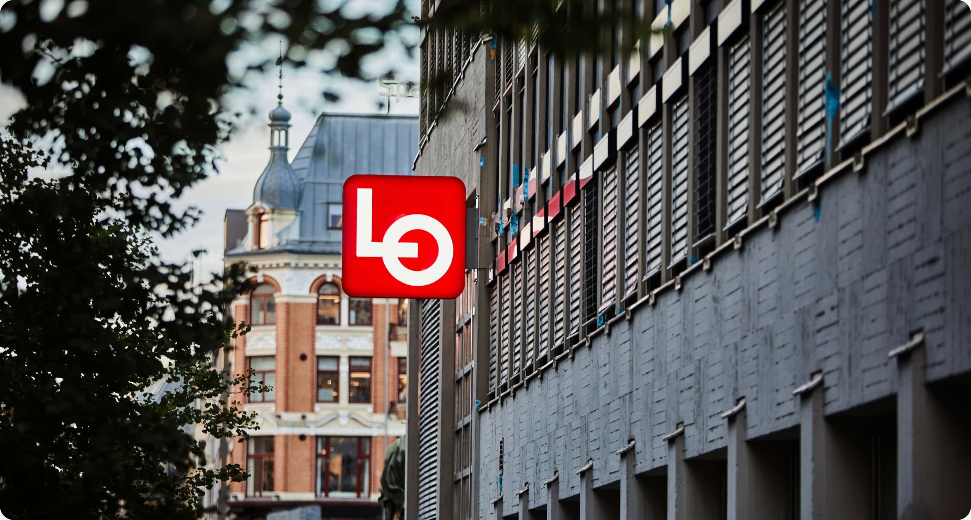 LOs røde logo på fasaden til LO-huset i Oslo, med den grå bygningen i forgrunn og et historisk murhus i bakgrunnen.