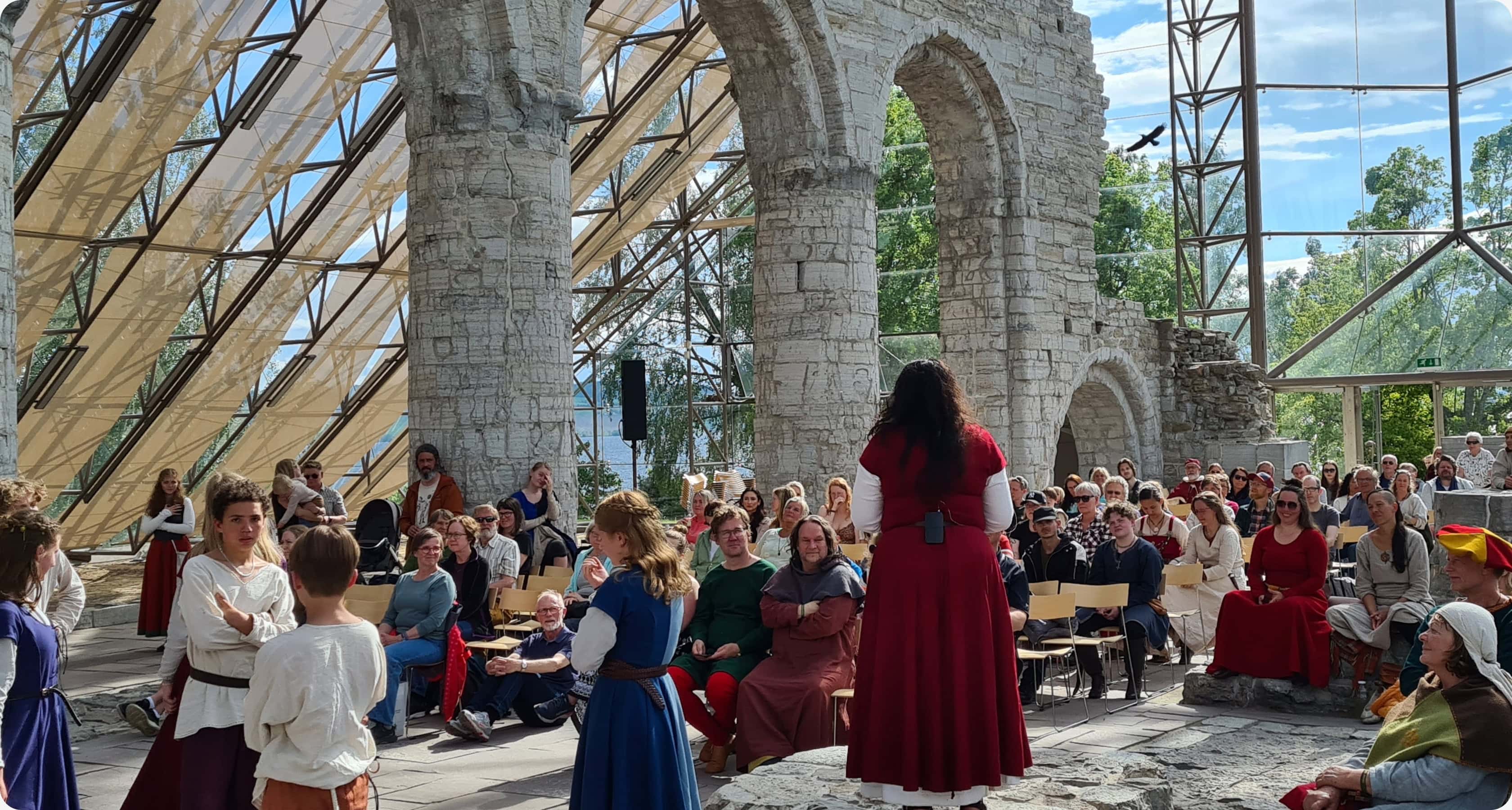Forestilling under Hamar Middelalderfestival i glassbygget på Domkirkeodden, med publikum og aktører i middelalderklær omgitt av ruiner og moderne konstruksjon.
