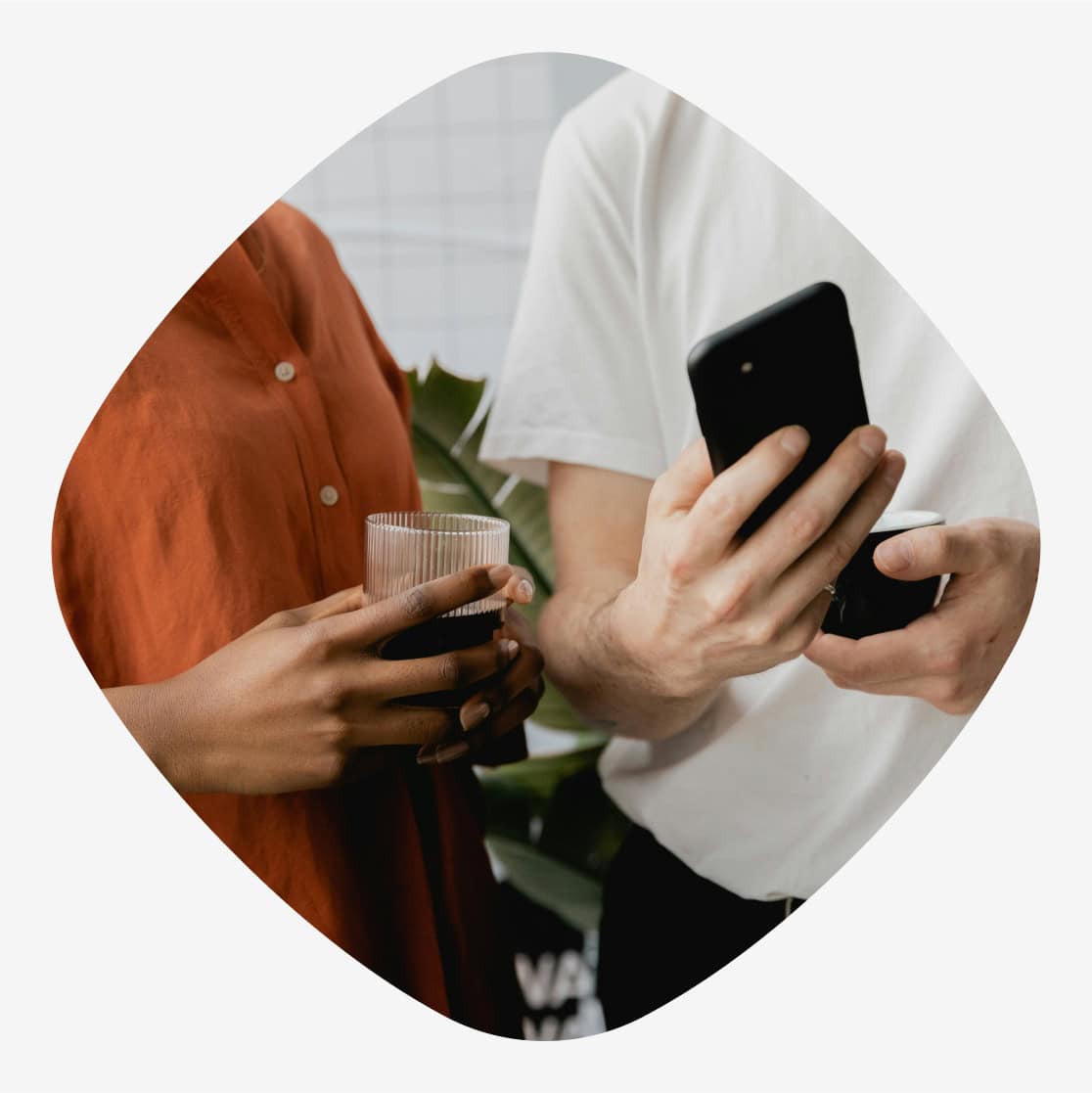 To personer står sammen, en i en rød skjorte og en i hvit t-skjorte, mens de holder en telefon og et glass.