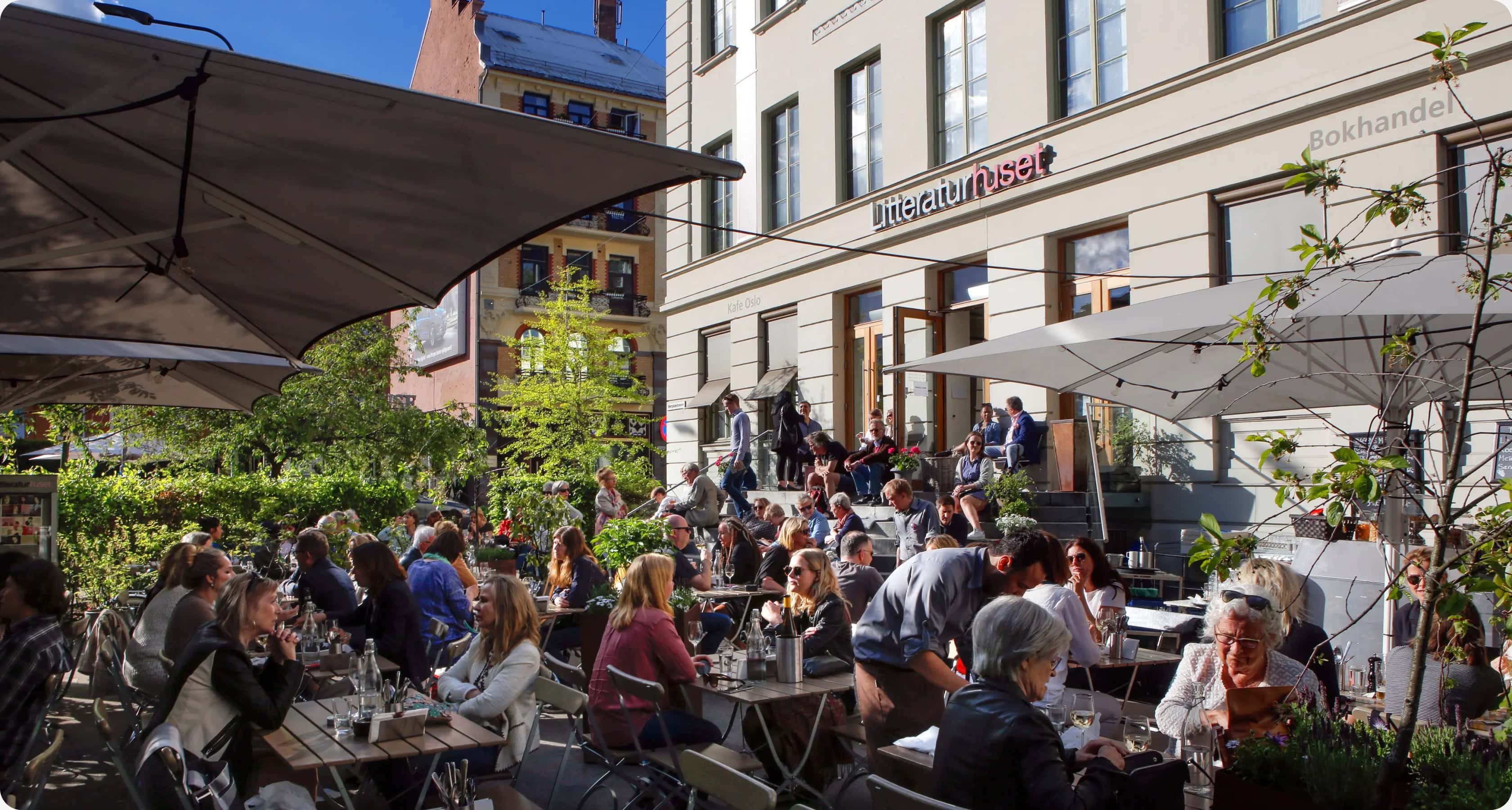 Uteserveringen ved Litteraturhuset i Oslo en solrik dag, med mange mennesker som sitter under parasoller og prater foran bygningen med skiltet «Litteraturhuset».