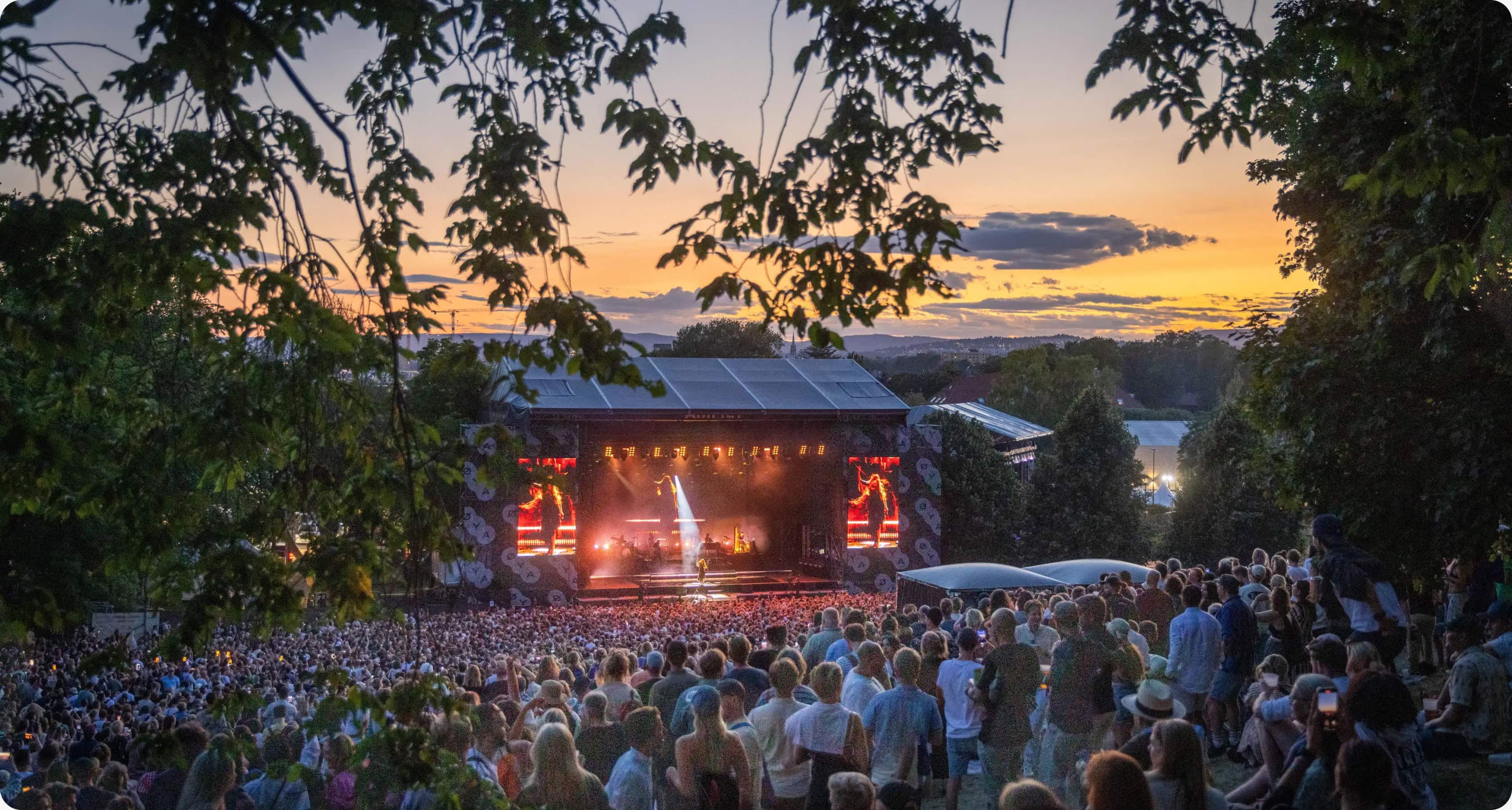 En stor folkemengde samlet foran en utendørsscene på Øyafestivalen i Oslo. Bildet er tatt fra en høyde med trær i forgrunnen, og scenen er opplyst av sterke lys og skjermer. Publikum står tett sammen mens solen går ned bak festivalområdet, og himmelen har en gyllen oransje og blå fargetone.