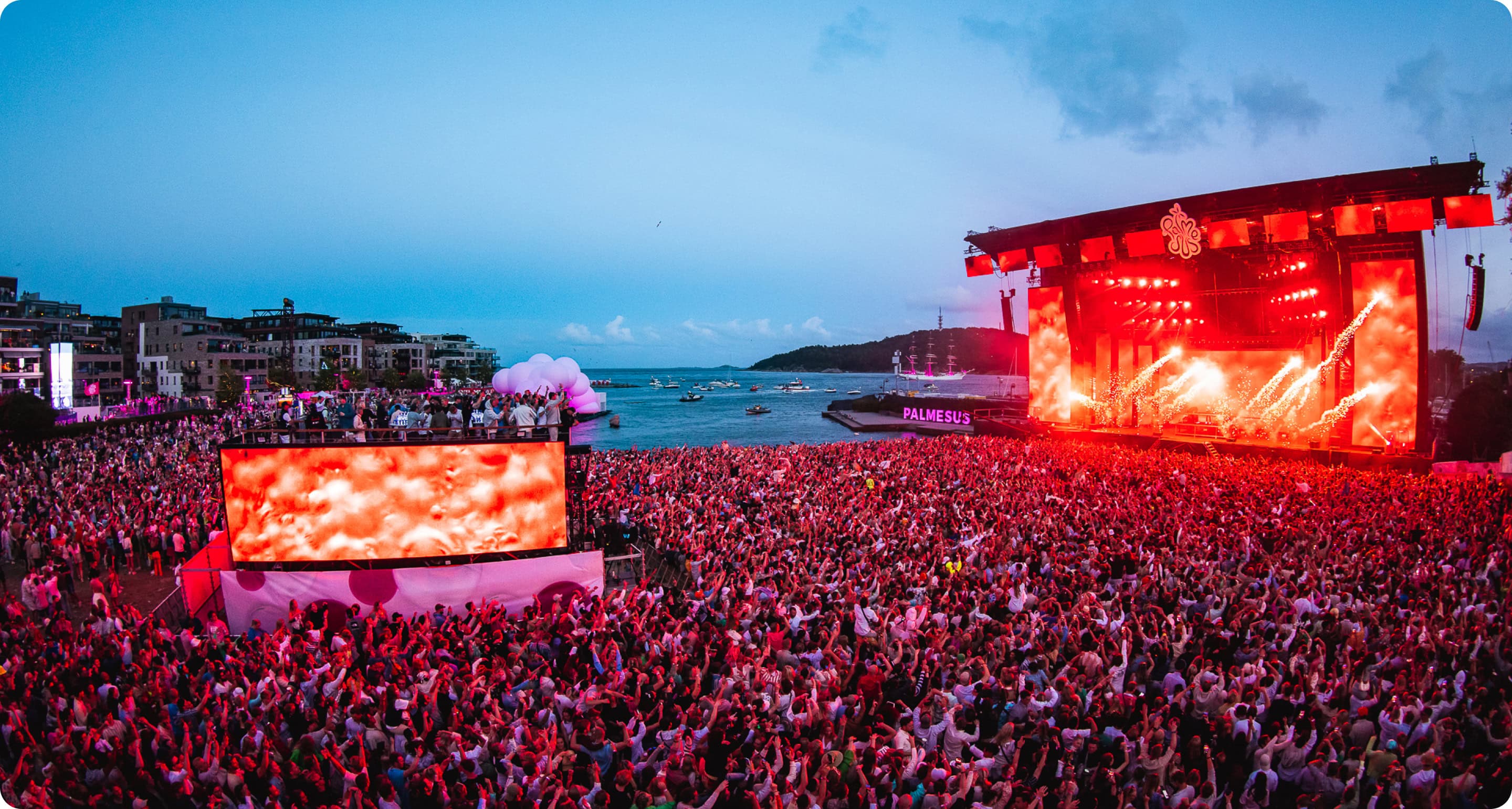 En folkemengde foran en scene med rødt lys på Palmesus-festivalen. Festivalområdet ligger ved sjøen, med båter ankret opp i bakgrunnen.