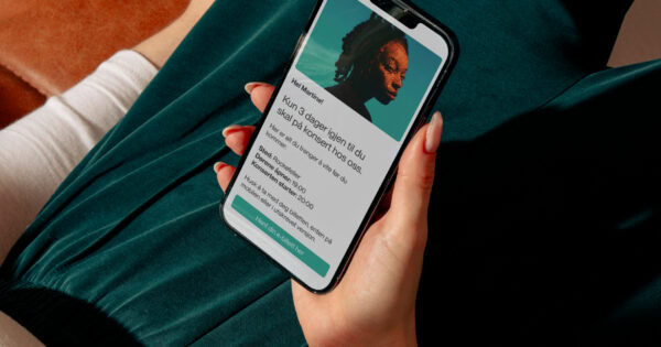 En person holder en mobiltelefon med en digital konsertbillett på skjermen, som viser konsertsted, tid og billettinformasjon.